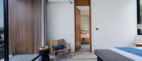 A modern bedroom with a large bed, a chair, a small table, and a view through a glass door.