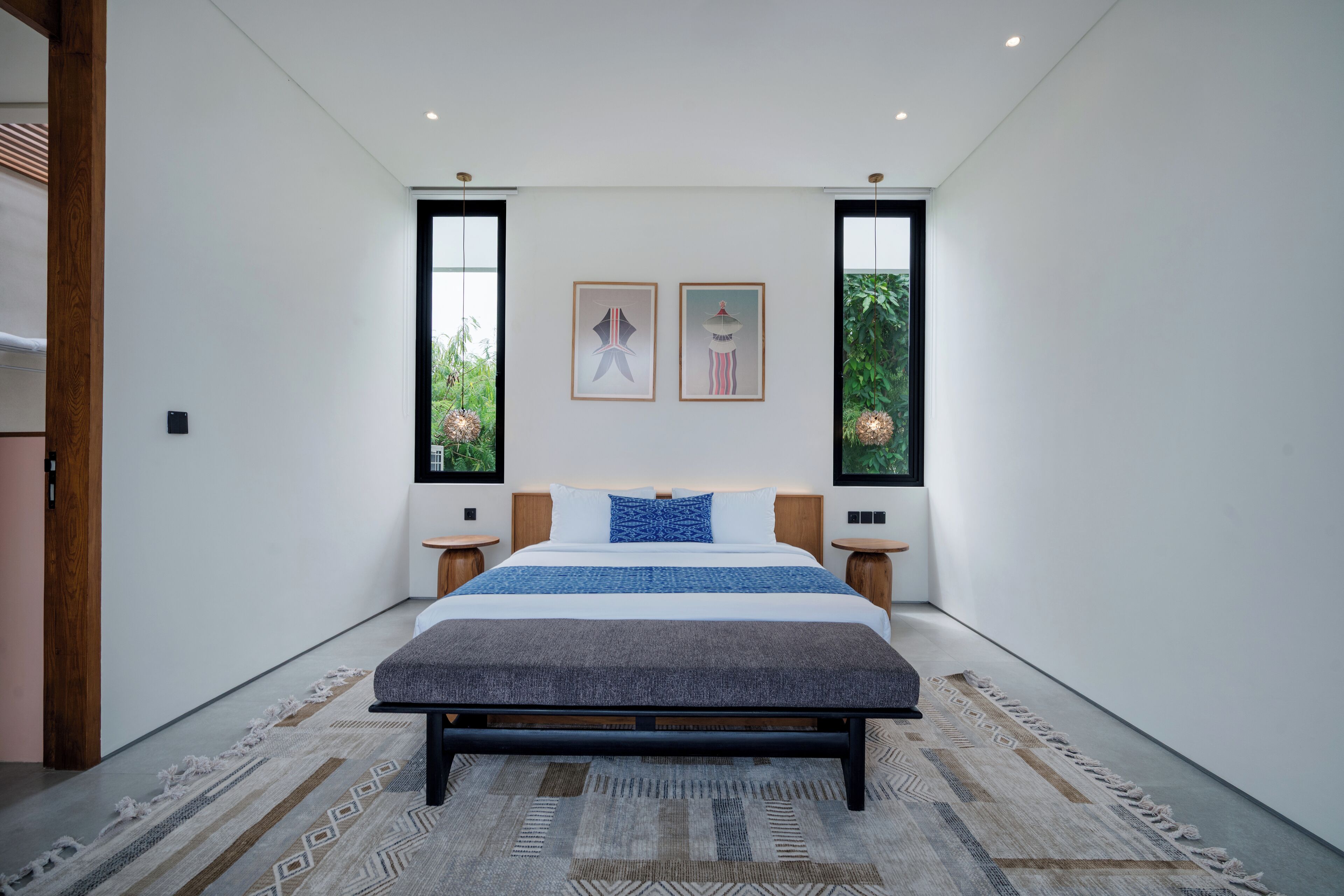 A modern bedroom with a large bed, two bedside tables, and a view of greenery through the windows.