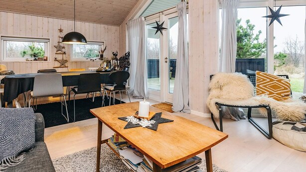 A cozy cabin interior with a dining area, a seating area with a sofa and armchair, and a large window with star decorations. - 5 Person Holiday Home in Brovst (Brovst)