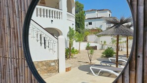 Blick durch ein rundes Fenster auf einen Ferienort mit Pool, Liegestühlen und einem Gebäude mit weißer Fassade und Bögen.