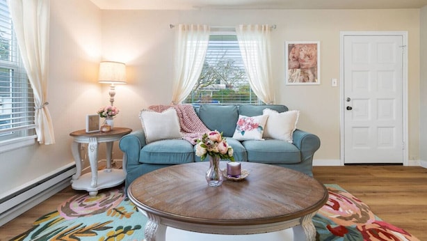 A cozy living room with a teal sofa, a round wooden coffee table, and a floral rug. There is a small side table with a lamp and a vase of flowers. A door and a window with curtains are visible in the background.