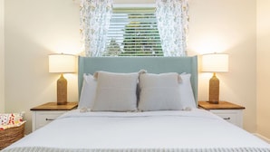 A neatly made bed with a light blue headboard, two bedside lamps, and a window with floral curtains.
