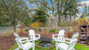 A patio area with Adirondack chairs and a round table, surrounded by trees and a wooden fence.