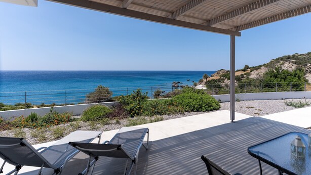Un espace extérieur abrité, doté de chaises longues et d’une table en verre, offrant une vue sur un paysage côtier avec une mer bleue cristalline et des collines.