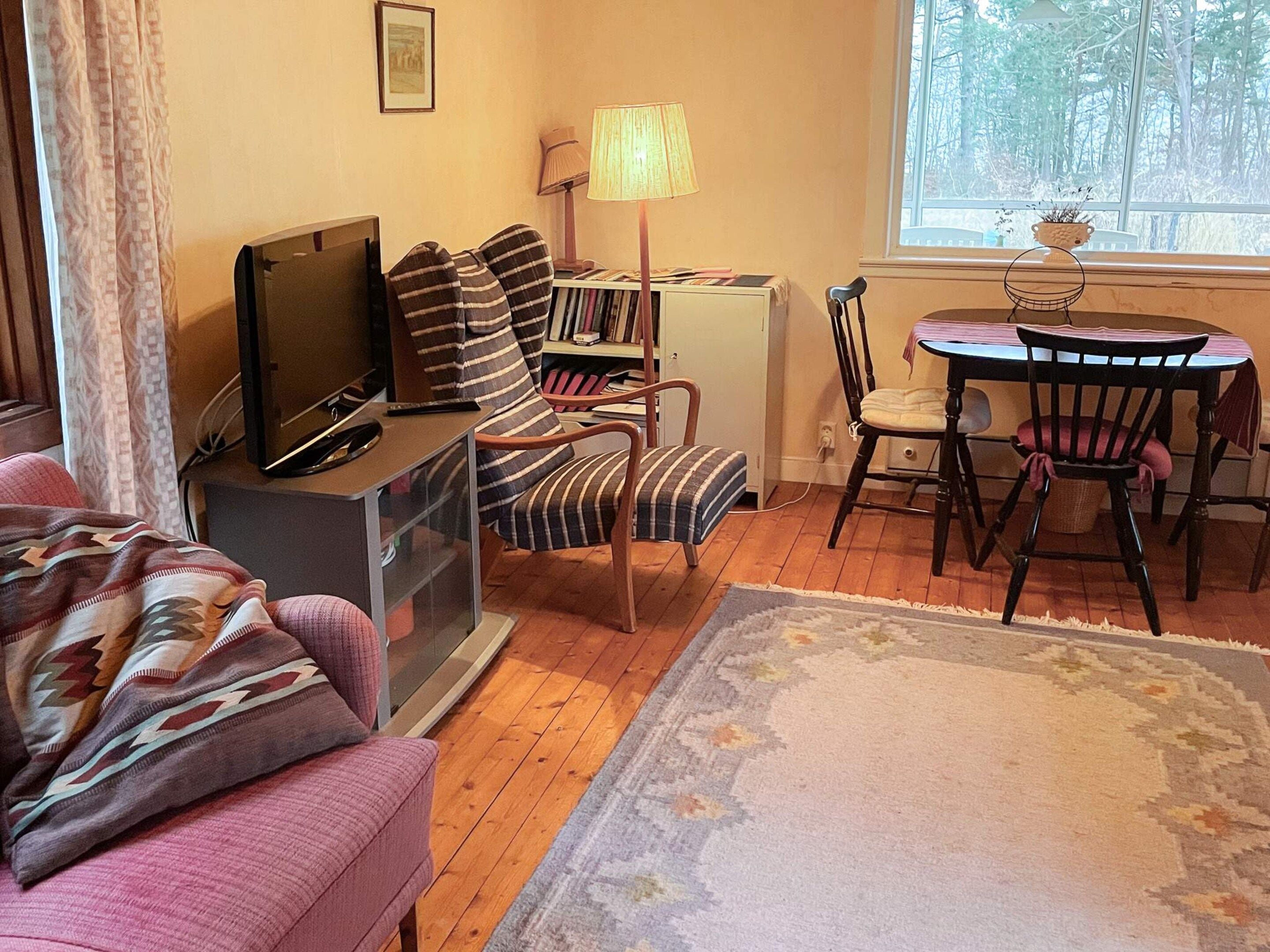 Un salon comprenant un canapé, un téléviseur, une bibliothèque, une lampe, une table à manger avec des chaises et une fenêtre donnant sur des arbres.