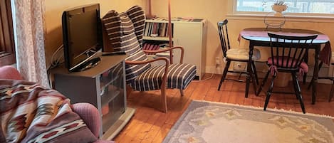 Un salon comprenant un canapé, un téléviseur, une bibliothèque, une lampe, une table à manger avec des chaises et une fenêtre donnant sur des arbres.