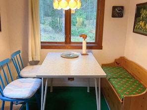 Ein Zimmer mit Tisch, Stühlen, einer Bank, einem Fenster mit Blick auf Grünflächen, einer Uhr und einem gerahmten Bild an der Wand.
