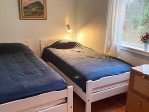 A bedroom with two single beds, a bedside table, a lamp, a window with curtains, and a painting on the wall.