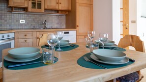 A dining table set with plates, glasses, and cutlery in a kitchen with wooden cabinets and a tiled backsplash.