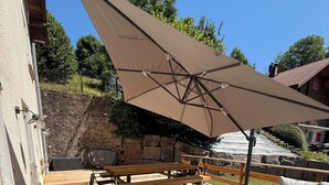 An outdoor patio with wooden flooring, a table with a cushion, a bench, and a large umbrella providing shade. - At 162 • Cosy 8-person house in the heart of the Vosges mountains (Anould)