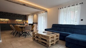 A modern living room with a blue sofa, a glass coffee table made from pallets, and a dining area with a table and chairs. - At 162 • Cosy 8-person house in the heart of the Vosges mountains (Anould)