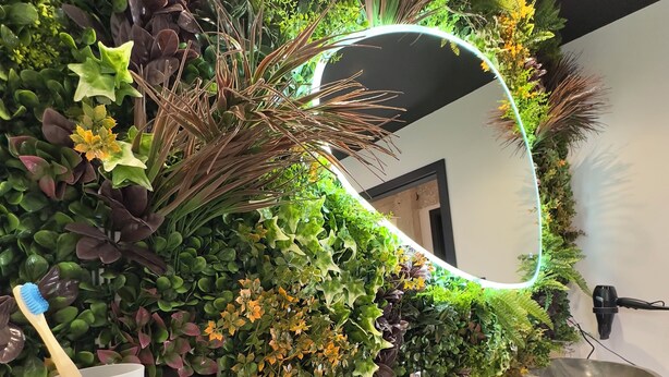 A bathroom with a wall-mounted mirror surrounded by lush green foliage, a wooden sink, and a toothbrush holder. - At 162 • Cosy 8-person house in the heart of the Vosges mountains (Anould)