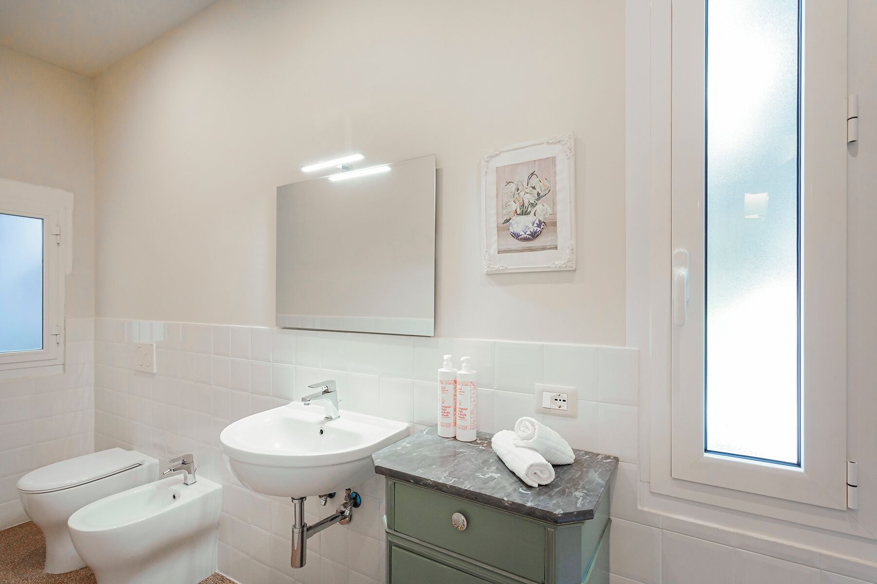 A bathroom with a white sink, a bidet, a mirror, and a window.