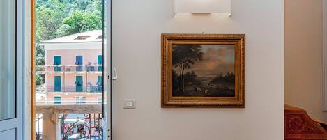A room with a framed painting on the wall, a light fixture above it, and a view of a building through an open door.