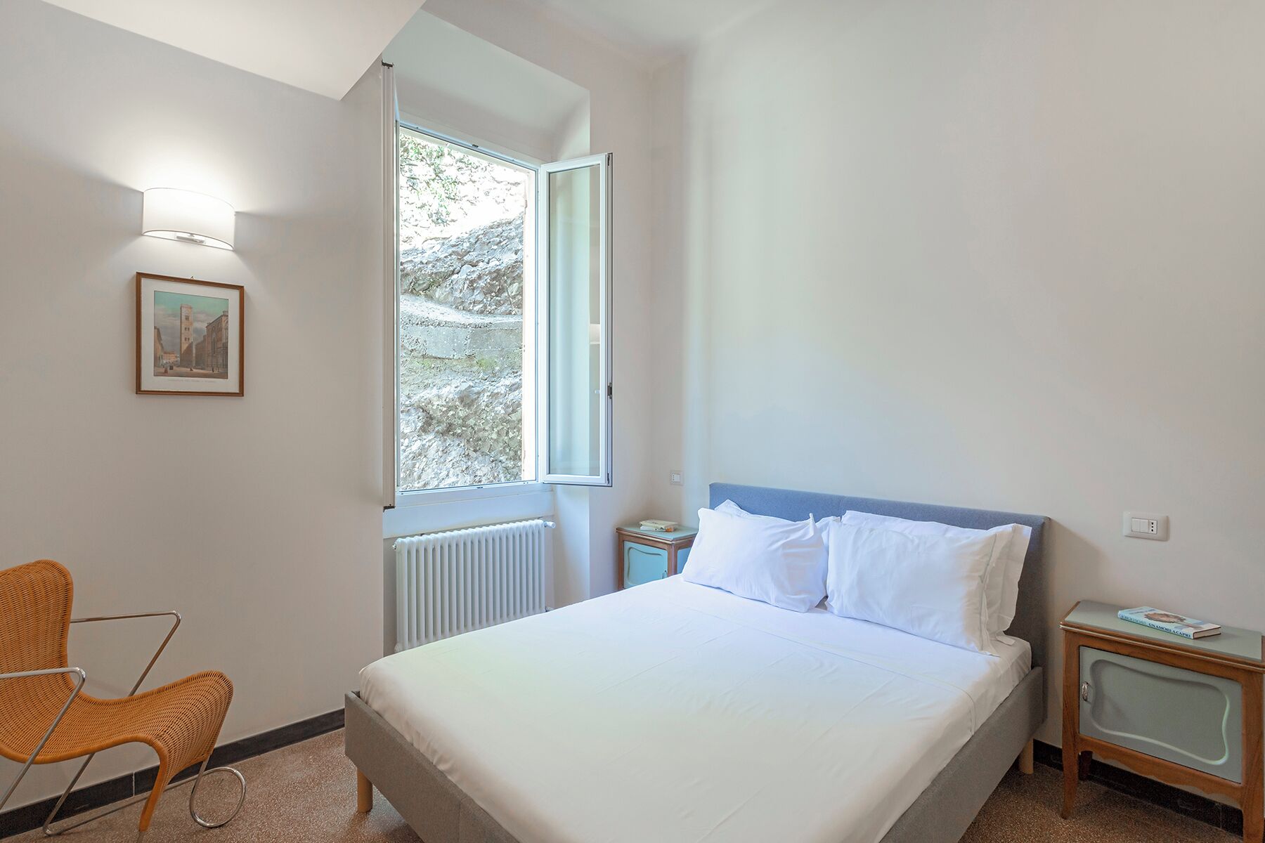 Ein Schlafzimmer mit Bett, Stuhl, Nachttisch, einem gerahmten Bild und einem Fenster mit Blick auf Bäume.