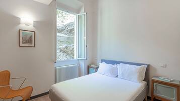 Ein Schlafzimmer mit Bett, Stuhl, Nachttisch, einem gerahmten Bild und einem Fenster mit Blick auf Bäume.