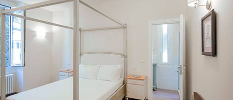 Un lit à baldaquin à quatre poteaux, avec du linge de lit blanc, une table de chevet, une petite commode et un tableau encadré au mur.