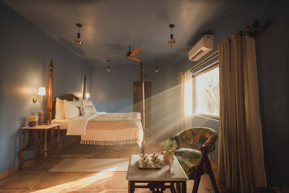 A bedroom with a bed, a chair, a table with jars, a standing lamp, and a ceiling fan.