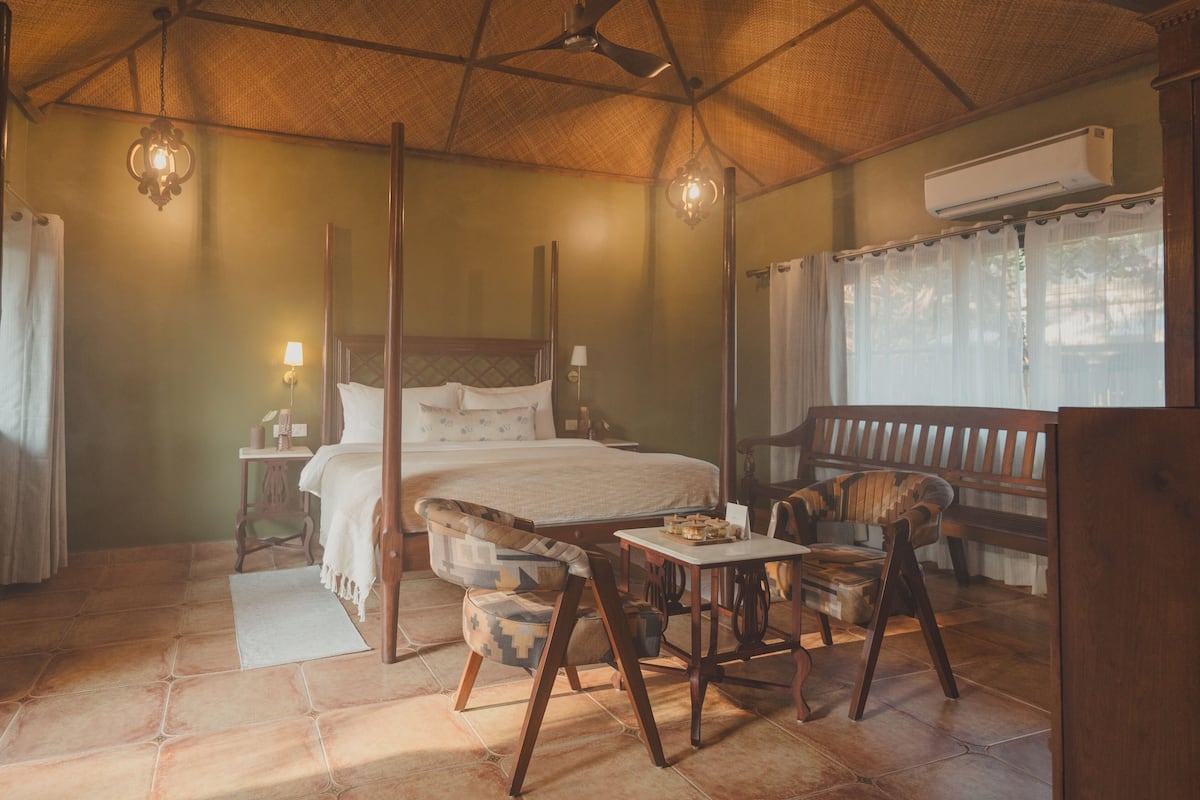 A spacious bedroom with a large bed, a wooden bench, a dining table, and a ceiling fan.