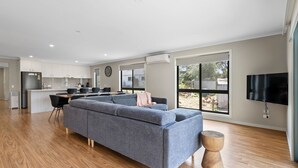 Een moderne woonkamer met een grijze bank, houten vloeren, een keuken met witte kasten en een groot raam met uitzicht op de achtertuin.