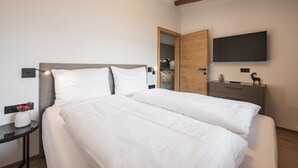 A hotel room with a bed, a bedside table with a vase of flowers, a TV mounted on the wall, and a wooden door leading to the bathroom. - Apartment in Kirchberg Near Schaterzlift (Kirchberg in Tirol)