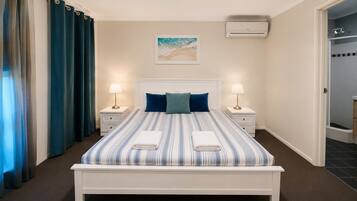 A hotel room with a bed, two bedside tables, a nightstand, a wall-mounted air conditioner, and a door leading to a shower area.