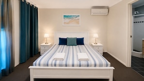 A hotel room with a bed, two bedside tables, a nightstand, a wall-mounted air conditioner, and a door leading to a shower area.