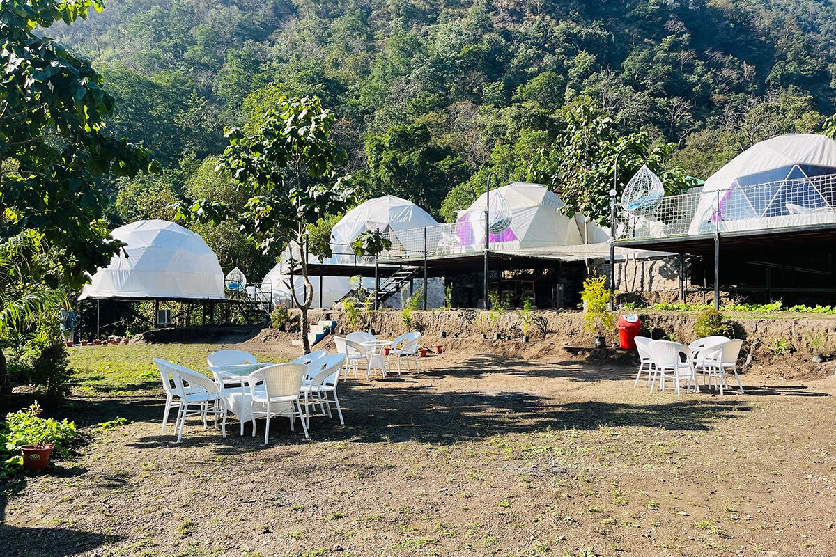 A campsite with dome-shaped tents, white plastic chairs, and tables arranged on a dirt ground.