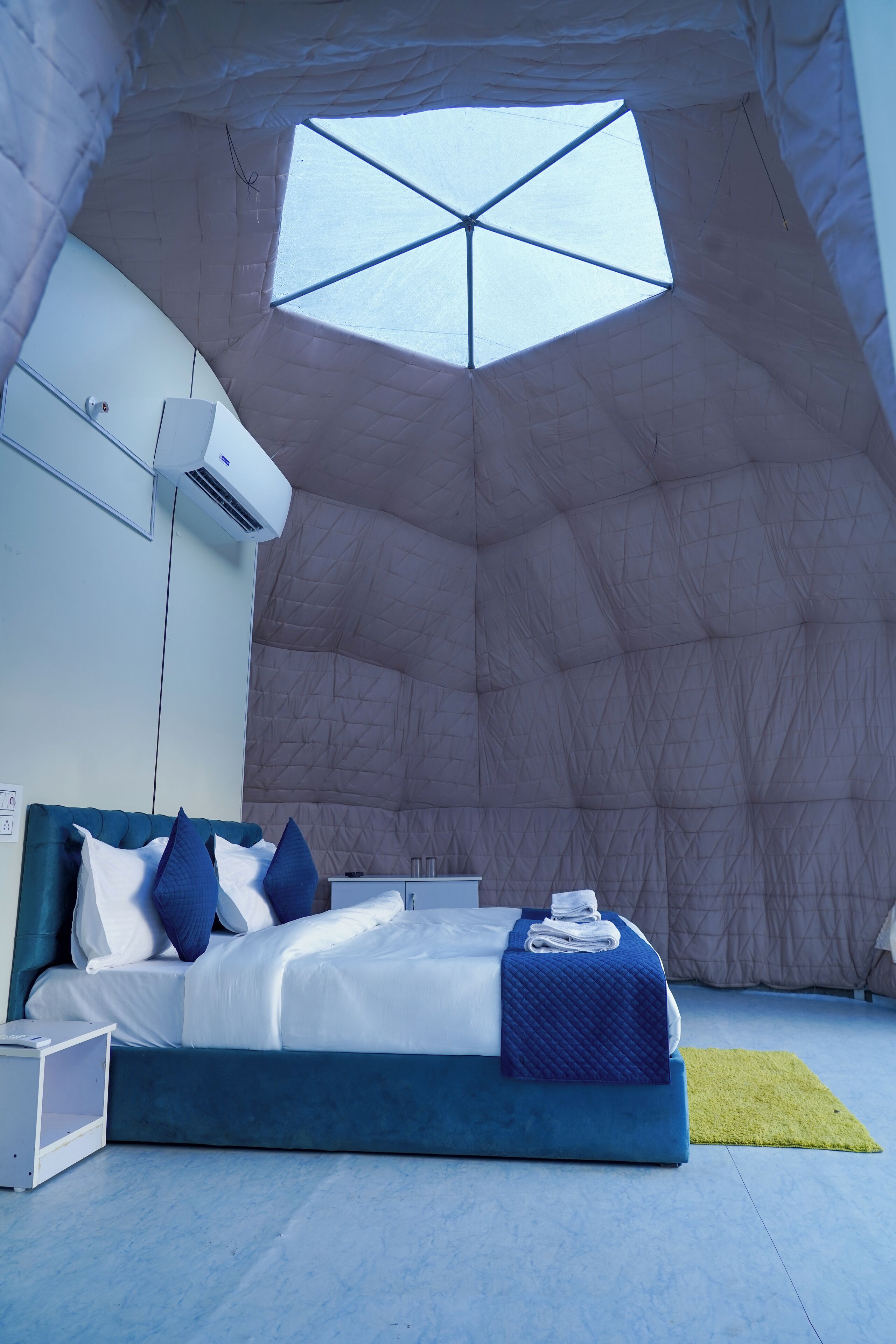 Ein Schlafzimmer mit einem Bett, Kissen, einem Nachttisch, einem kleinen Beistelltisch und einem Fenster mit geometrischer Aussicht.