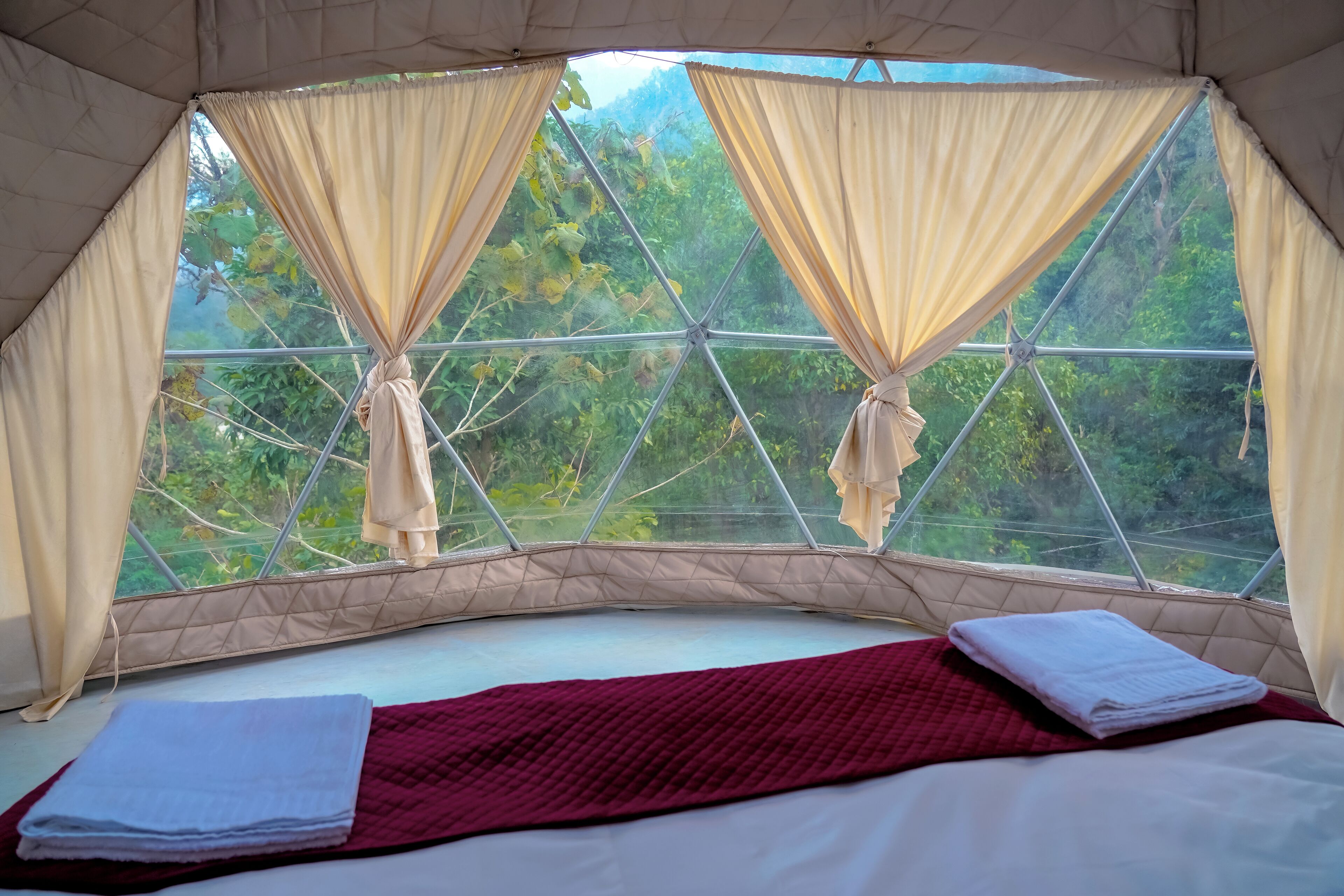 A tented room with a bed, two windows with curtains, and a view of greenery.