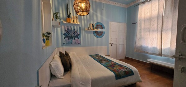 A bedroom with a bed, a bedside table, a wall-mounted shelf with decorative items, a door, and a window with curtains. - The Ganges By Boho Nomad Nook (Rishikesh)