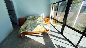 A bedroom with a bed, wooden nightstands, a large window, and a tiled floor.