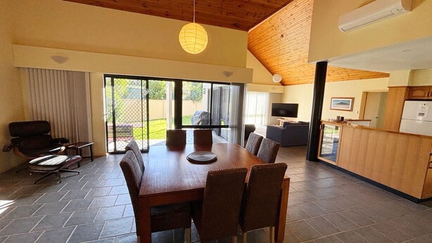 A spacious dining area with a large table and chairs, a kitchen area with a refrigerator and a flat-screen TV, and a sliding glass door leading to an outdoor patio.