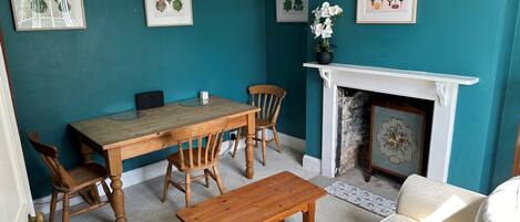 A dining area with a wooden table and chairs, a white sofa, a fireplace, and framed botanical prints on teal walls.