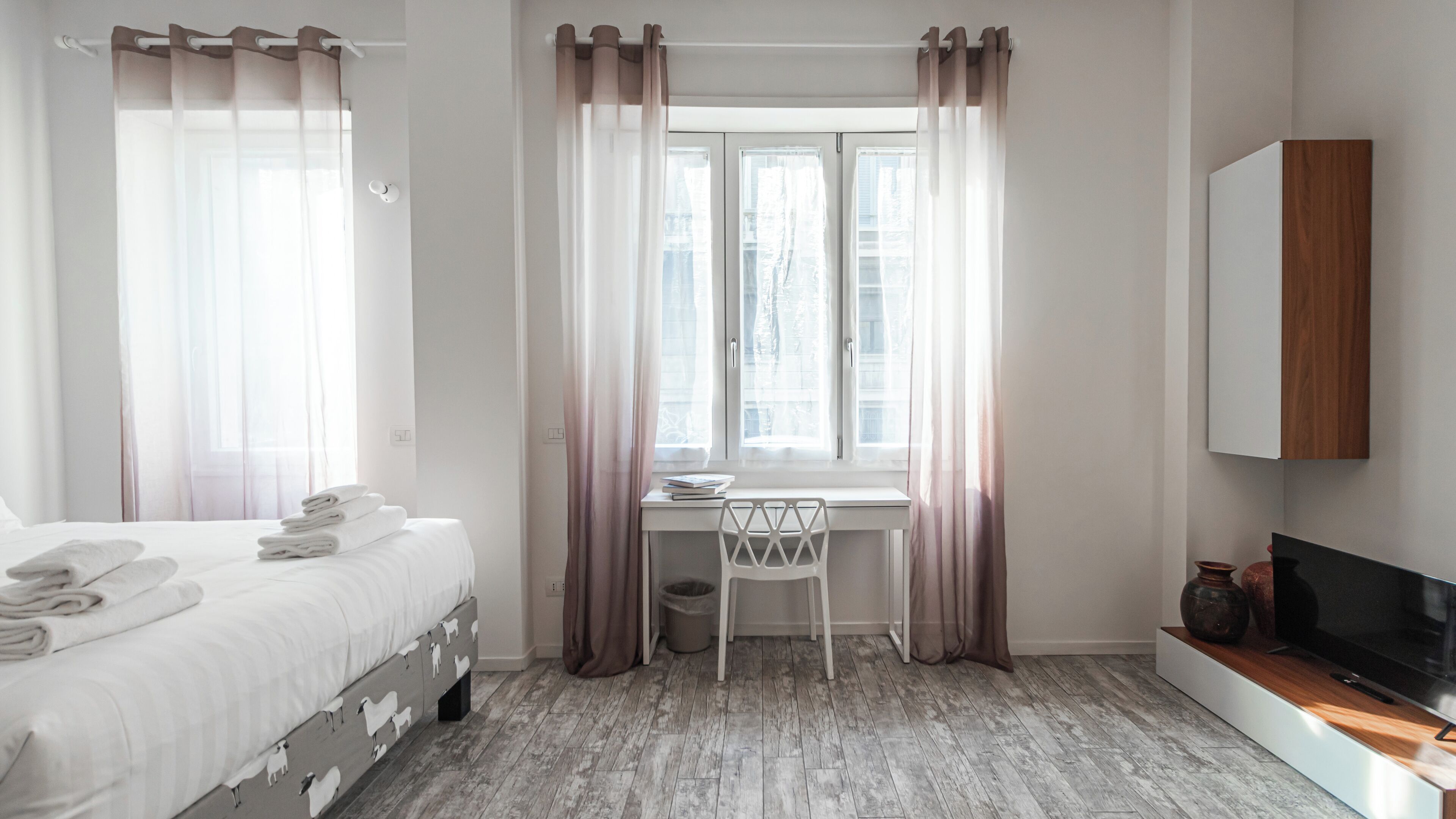 A hotel room with a bed, a desk, a chair, a TV, and a window with curtains.