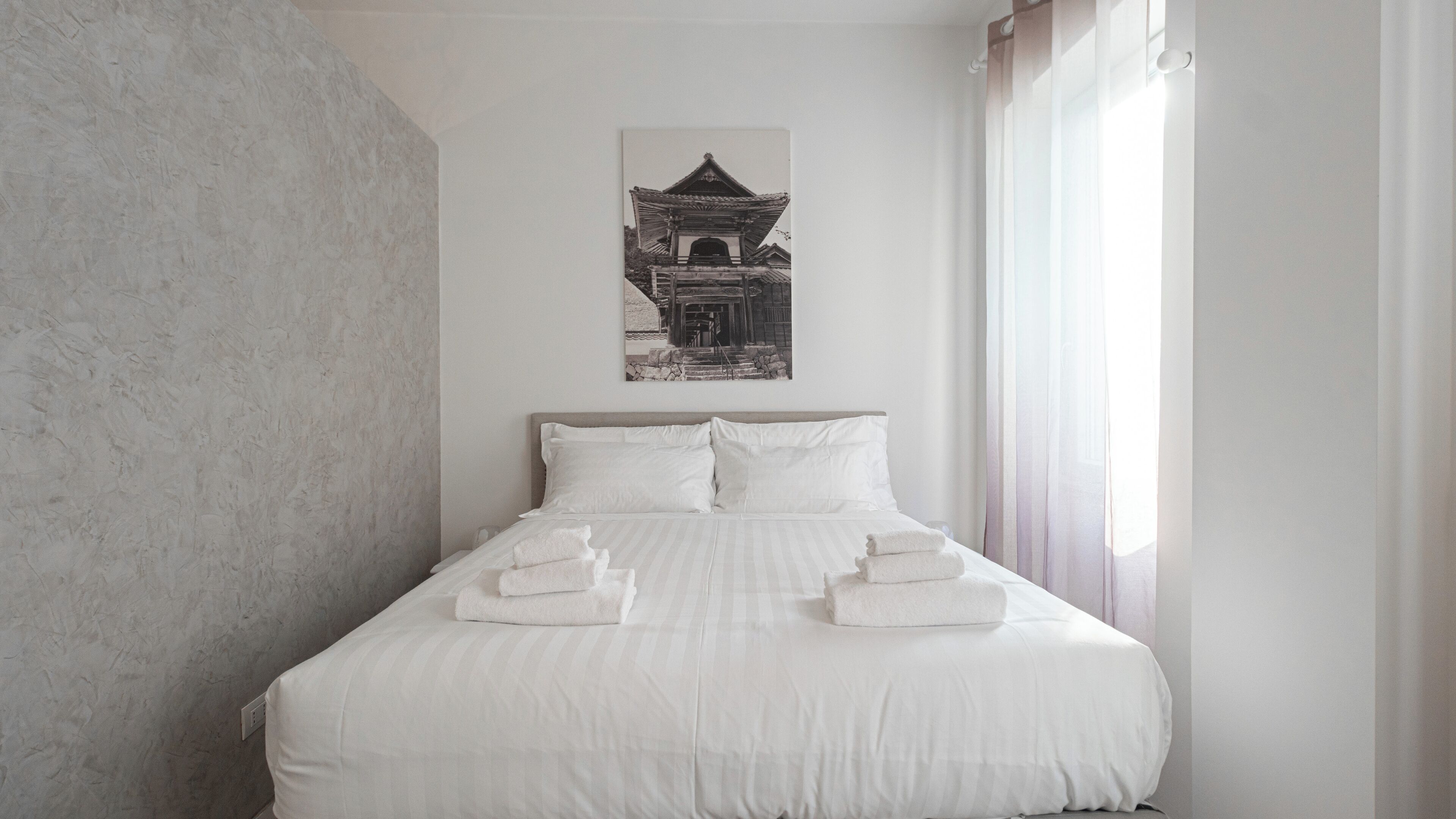 A neatly made bed with white linens and towels, a headboard, and a wall-mounted picture of a traditional building.