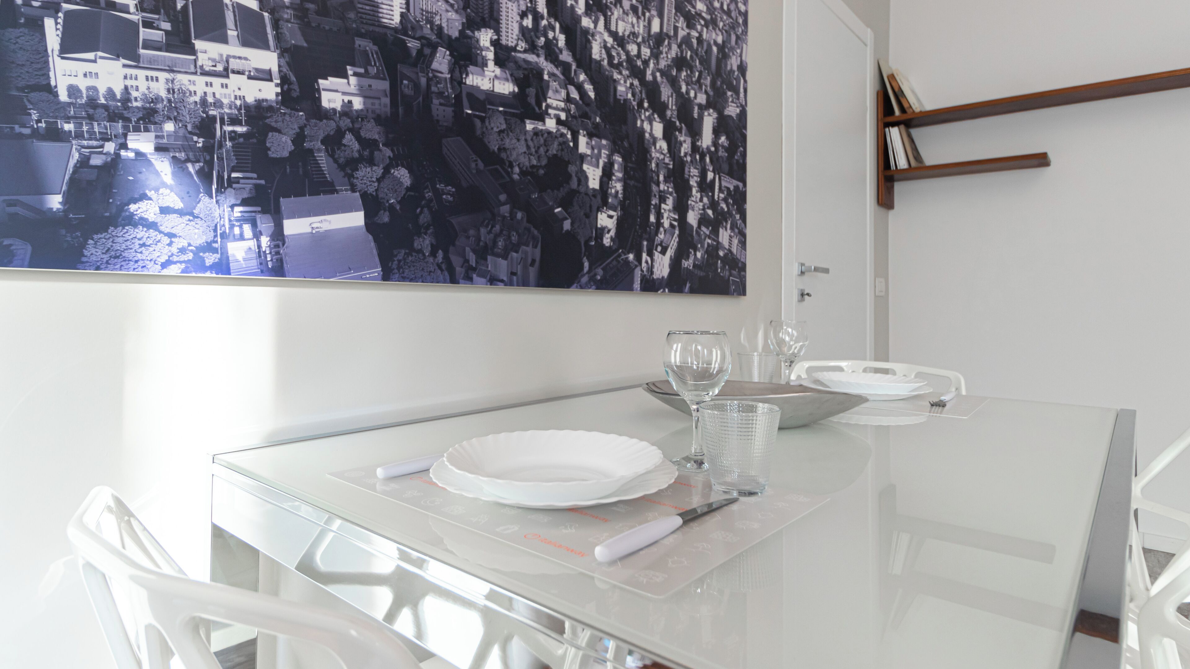 Una mesa de comedor dispuesta con platos blancos, vasos y cubiertos, con un cuadro de un paisaje urbano en la pared superior.