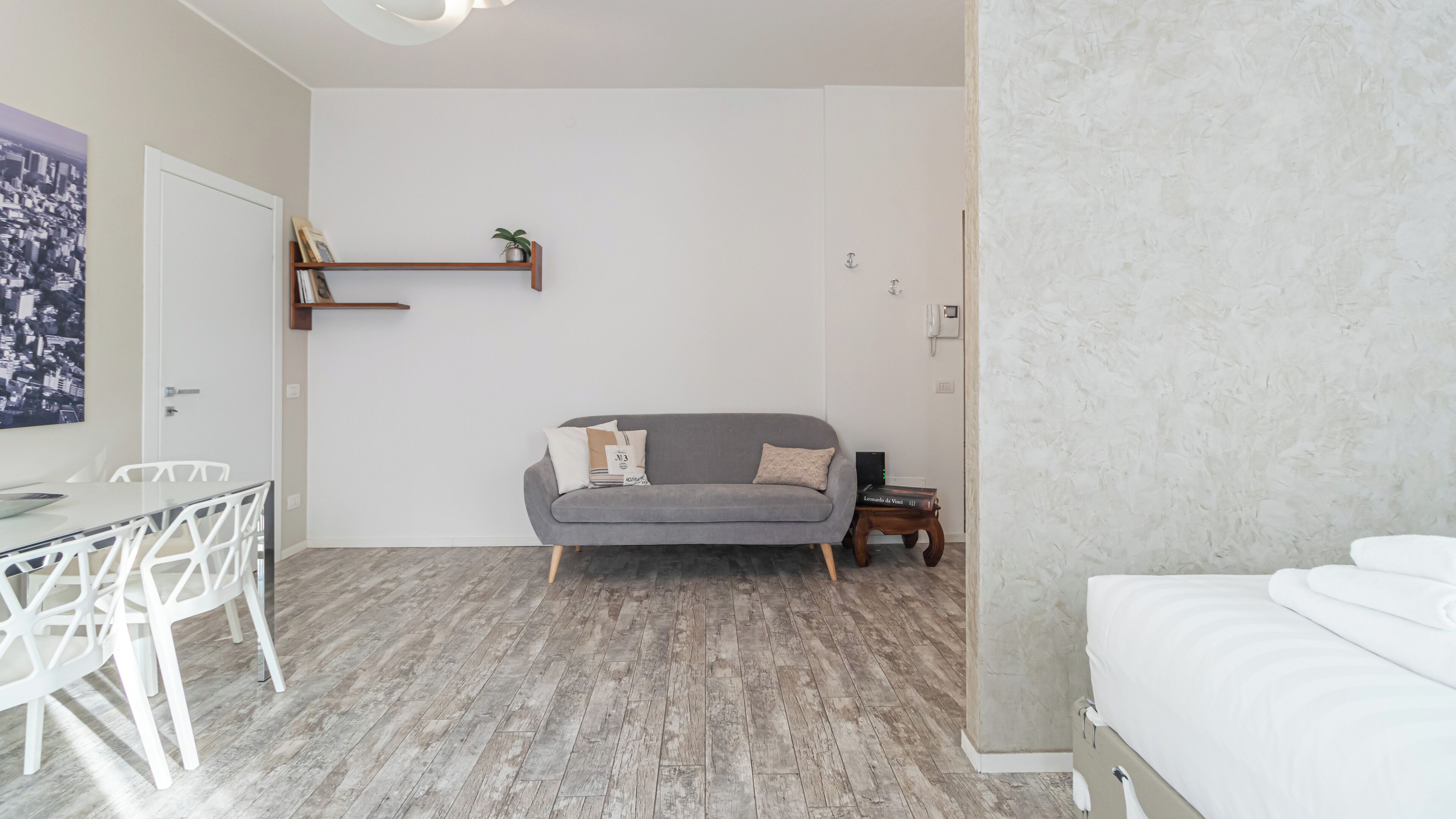 A modern, well-lit room with a grey sofa, a small wooden side table, a white bed, and a dining area with a glass table and white chairs.