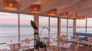 Ein Restaurant direkt am Strand mit Korbmöbeln, großen Fenstern und hängenden Lampen aus Korbgeflecht.