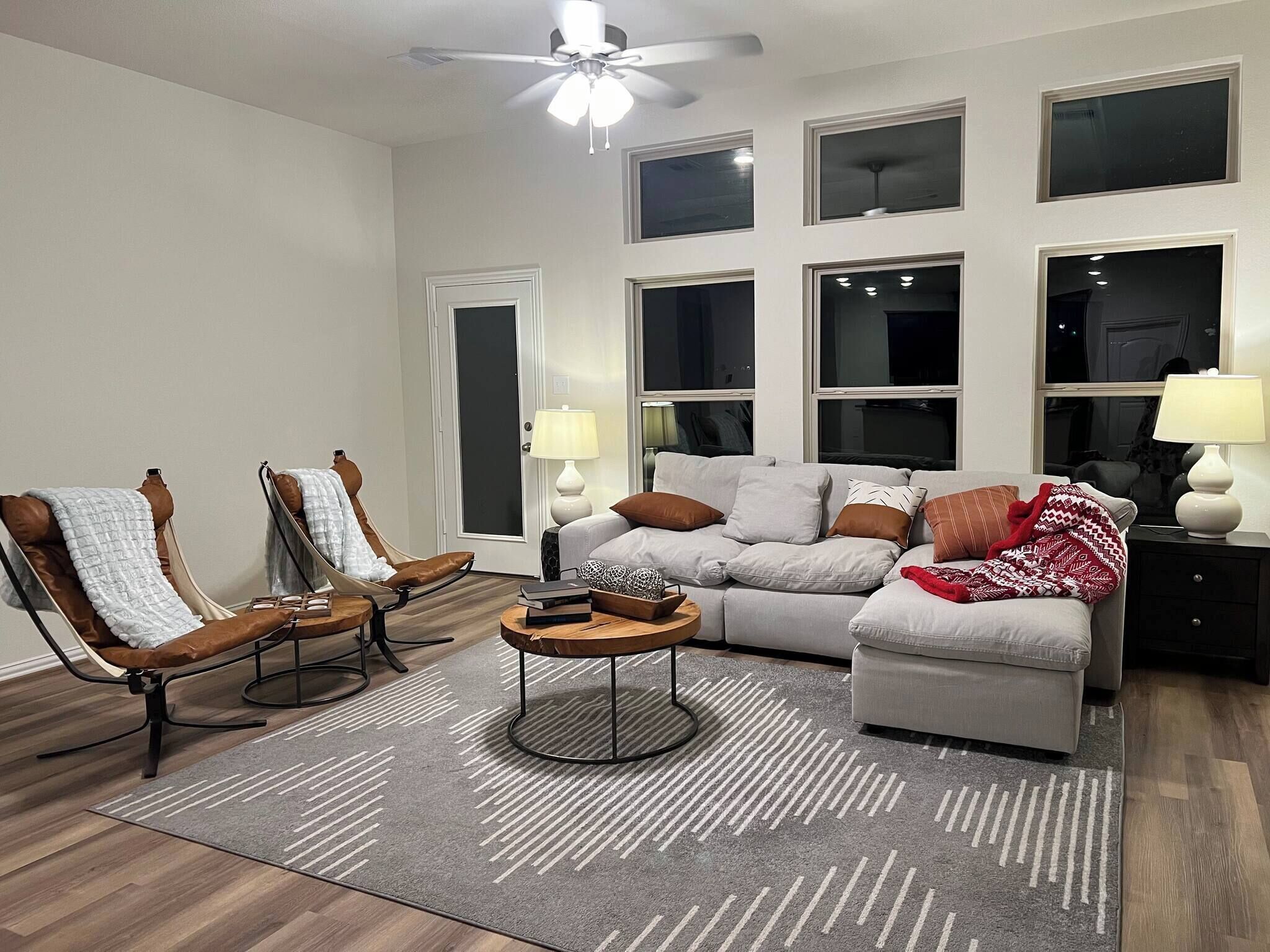 A living room with a grey sofa, two armchairs, a coffee table, and a rug. There are multiple windows and a ceiling fan.