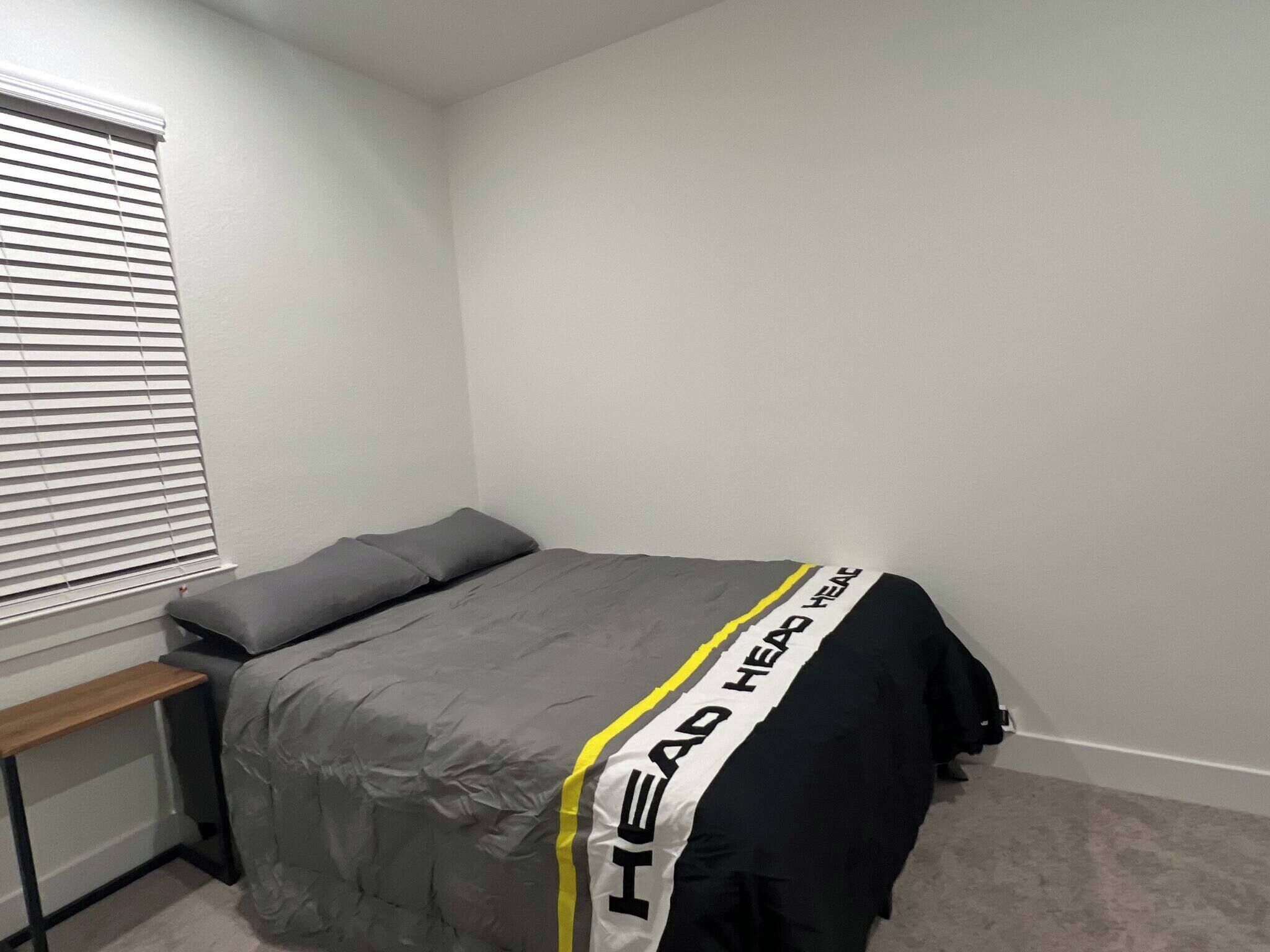 A bedroom with a bed, a bedside table, a window with blinds, and a wall with a partial view of a door.