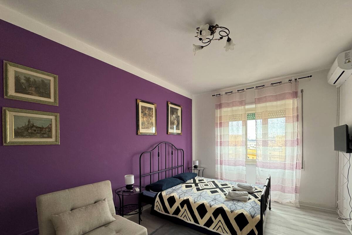 Slaapkamer met paarse muren, een bed met een beddekleed met dessin, een stoel, een kleine tafel met een lamp, en twee ingelijste foto’s aan de muur.