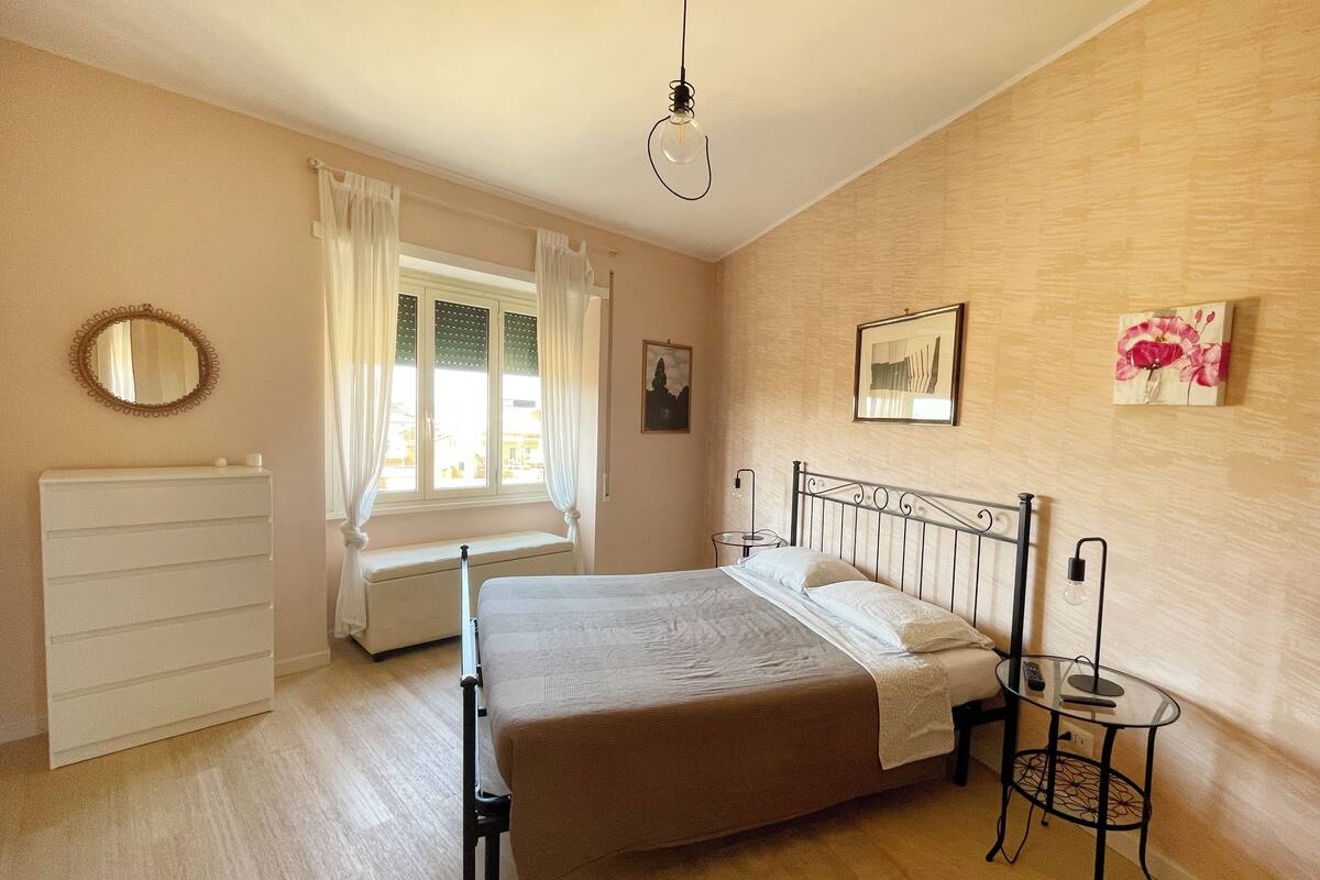 Een slaapkamer met een bed, nachtkastje, lamp, raam met gordijnen, radiator en wanddecoratie.