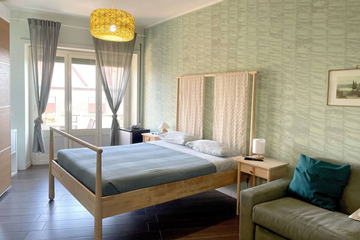Een slaapkamer met een houten bed, een nachtkastje met lamp, een groene bank en een raam met gordijnen.