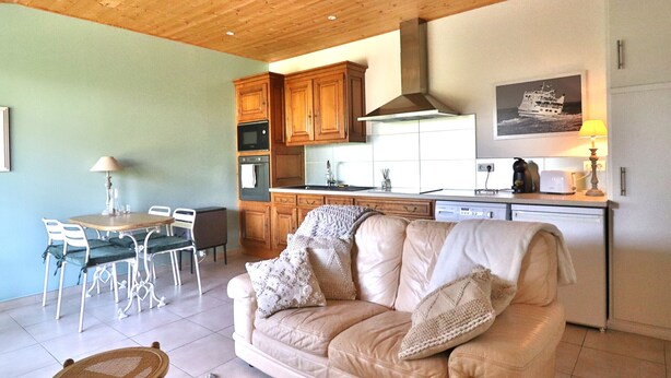 A cozy living room with a beige sofa, a dining table with chairs, and a kitchen area with wooden cabinets and appliances. - Apartment with private garden and modern amenities (Groix)