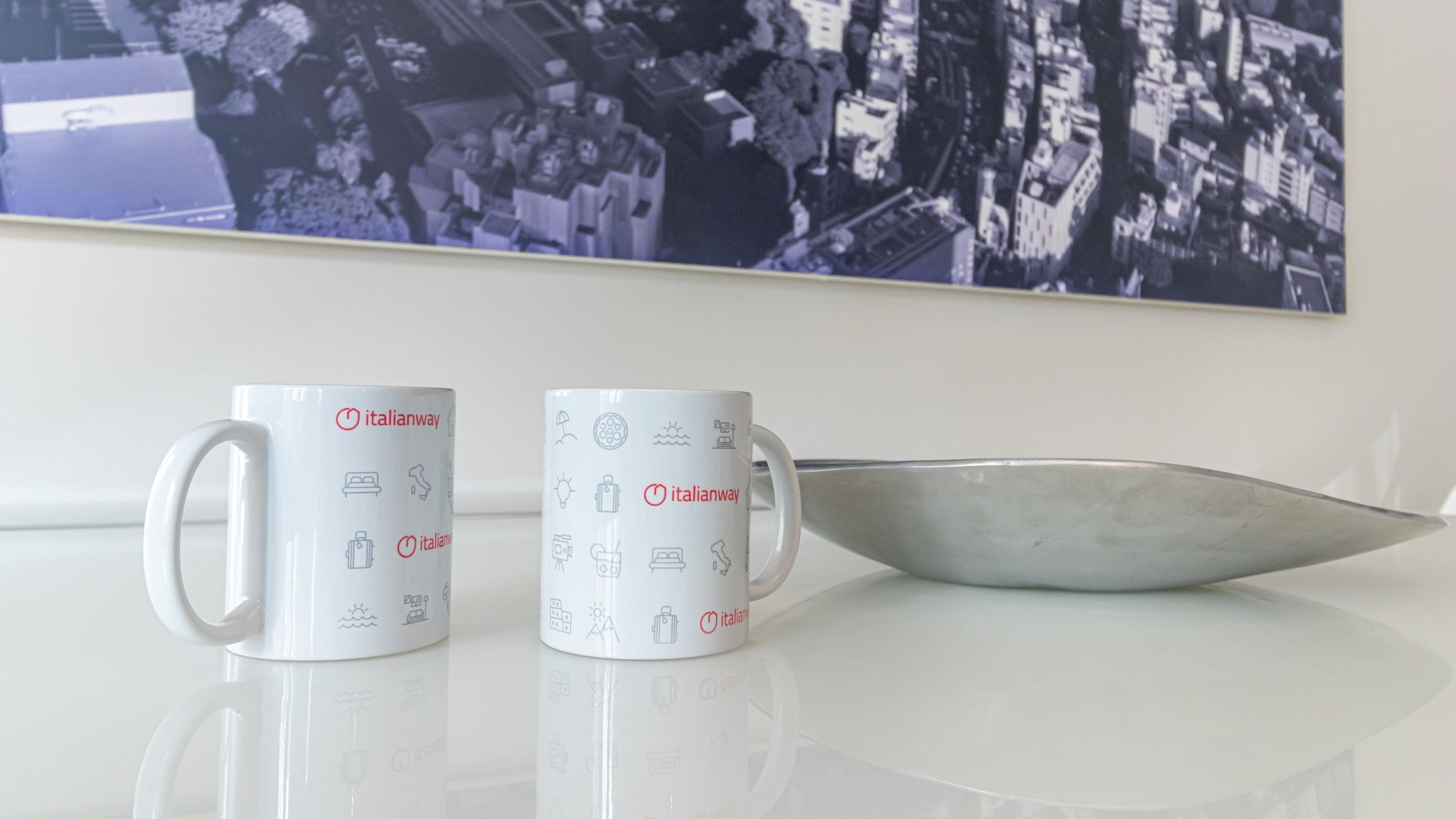 Dos tazas blancas con iconos y texto relacionados con viajes, y un bol metálico sobre una superficie reflectante.