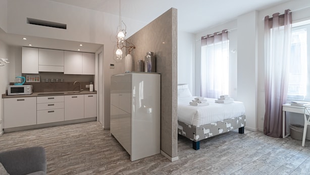 Ein modernes Hotelzimmer mit einer Küchenzeile, einem Bett mit gemustertem Kopfteil und einem großen Fenster mit transparenten Vorhängen.
