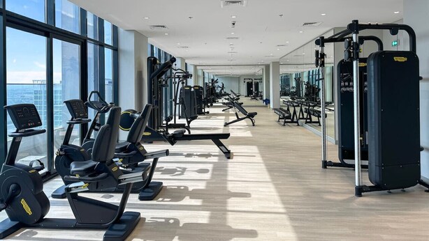 A modern gym with cardio machines, weightlifting equipment, and large windows offering city views. - Sleek Studio Apartment in Azizi Riviera- Suiteable (Dubai)