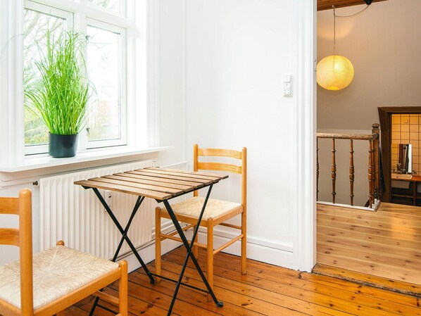 A room with a wooden floor, a window with a potted plant, two wooden chairs, a small table, and a hanging light fixture. - Apartment in Ringkobing With Garden (Ringkobing)