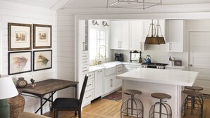 Een moderne keuken met witte kastjes, een centraal kookeiland met krukken, een eettafel met stoelen en kunstwerken in lijsten aan de muur.